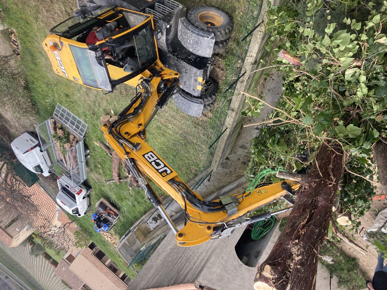 Tronc en cours d’abattage près d’une habitation, avec un engin et du bois coupé